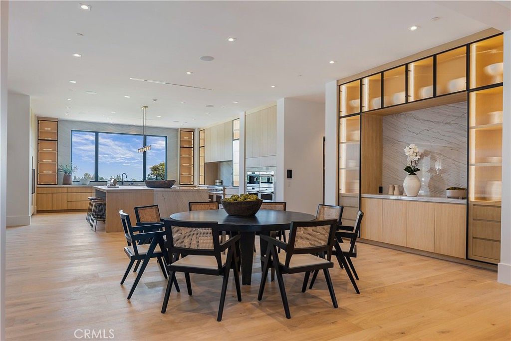 This expansive, open-concept kitchen and dining area features a sophisticated blend of light wood cabinetry, sleek marble-like backsplashes, and high-end stainless steel appliances. A large central island with bar seating complements the circular dining table in the foreground, creating a seamless flow for entertaining. The space is bathed in natural light from a large window, highlighting the warm hardwood floors and modern, minimalist aesthetic.
