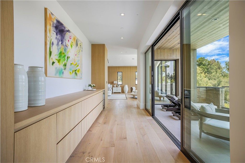 This elegant hallway features light-toned hardwood flooring and a long, custom built-in wooden console that provides a clean, minimalist aesthetic. Large floor-to-ceiling sliding glass doors open onto a covered patio, seamlessly blending indoor and outdoor living spaces while flooding the area with natural light. The perspective leads the eye through the hallway toward a bright, airy bedroom suite in the background, emphasizing the home's open and sophisticated architectural design.