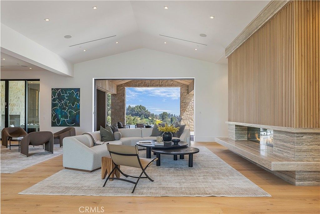 This spacious, modern living room features a high vaulted ceiling and a striking floor-to-ceiling fireplace with a wood-slat accent wall. A large, plush sectional sofa centers the room, complemented by a layered coffee table and a neutral area rug, all set against light hardwood floors. The room is bathed in natural light from a large opening that frames a scenic outdoor view, creating an airy and sophisticated atmosphere.