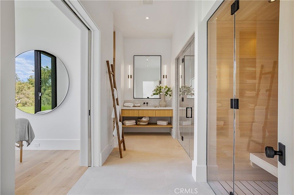 This luxurious primary bathroom features a clean, modern aesthetic with a floating wood vanity, a minimalist mirror, and a light-toned color palette. To the right, a glass-enclosed sauna adds a spa-like element, while a wooden ladder towel rack leans against the wall, enhancing the organic, contemporary design. The perspective looks down a bright, airy hallway that connects the vanity area to the rest of the suite, creating a seamless and sophisticated flow.