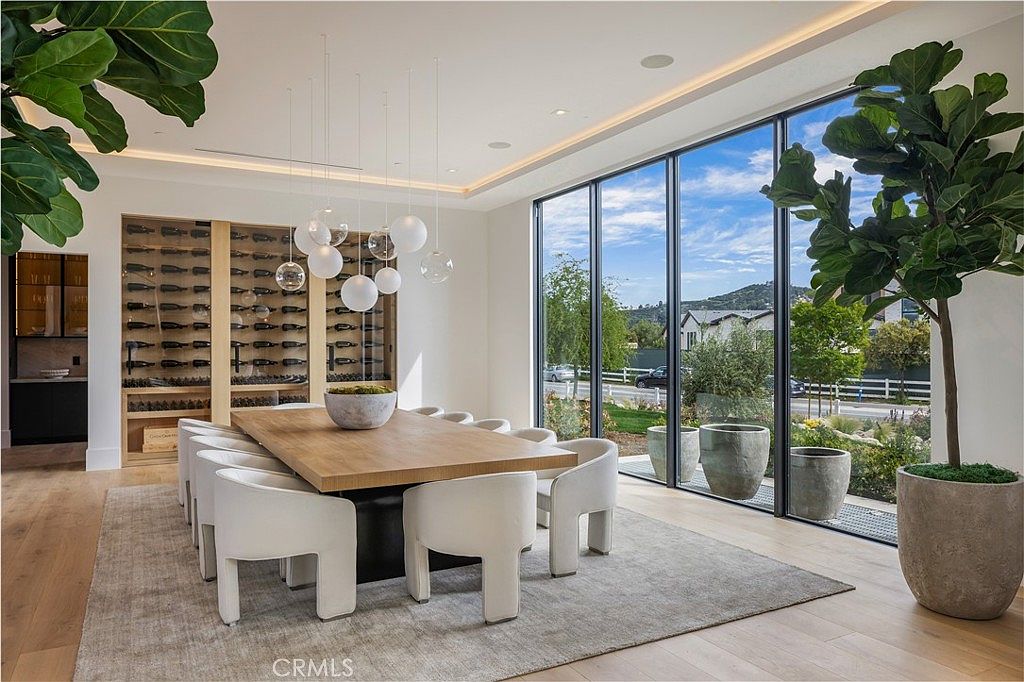 This sophisticated dining room features a large, light-wood table surrounded by modern, curved white chairs, set atop a textured area rug. A striking cluster of spherical pendant lights hangs above the table, while a floor-to-ceiling glass wall provides an expansive view of the outdoor landscape. To the side, a built-in wine display adds a touch of luxury, creating an elegant and airy atmosphere perfect for formal entertaining.