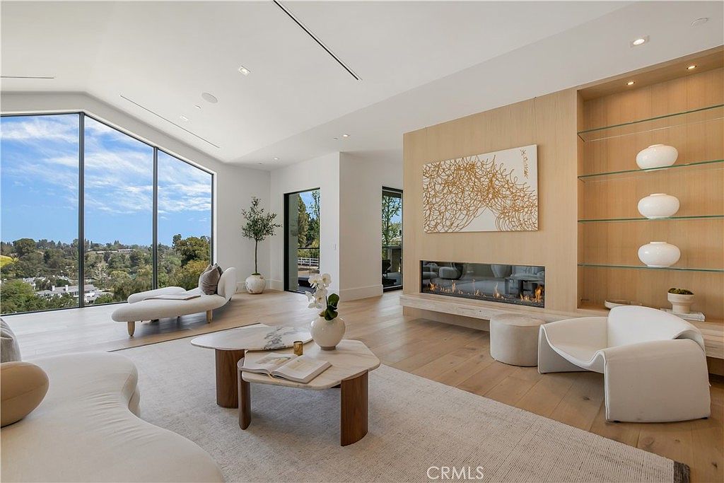 This bright and airy living room features a contemporary design with floor-to-ceiling windows that offer expansive views of the surrounding landscape. The space is anchored by a sleek, linear fireplace set into a light wood-paneled wall, complemented by minimalist furniture and a neutral color palette. The open-concept layout creates a sophisticated and tranquil atmosphere, perfect for modern luxury living.