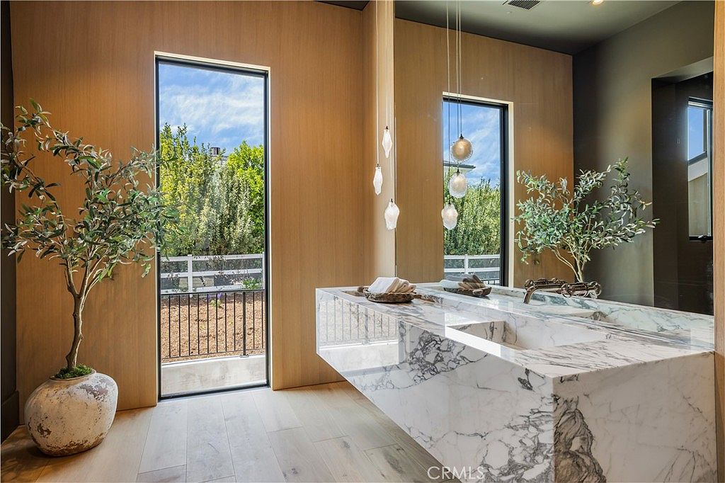 This luxurious primary bathroom features a striking floating marble vanity with dramatic veining, complemented by warm wood-paneled walls and light hardwood flooring. A large mirror reflects the space, while elegant pendant lights hang above the sink, and a floor-to-ceiling window provides a serene view of the outdoor greenery. The overall aesthetic is sophisticated, modern, and spa-like.