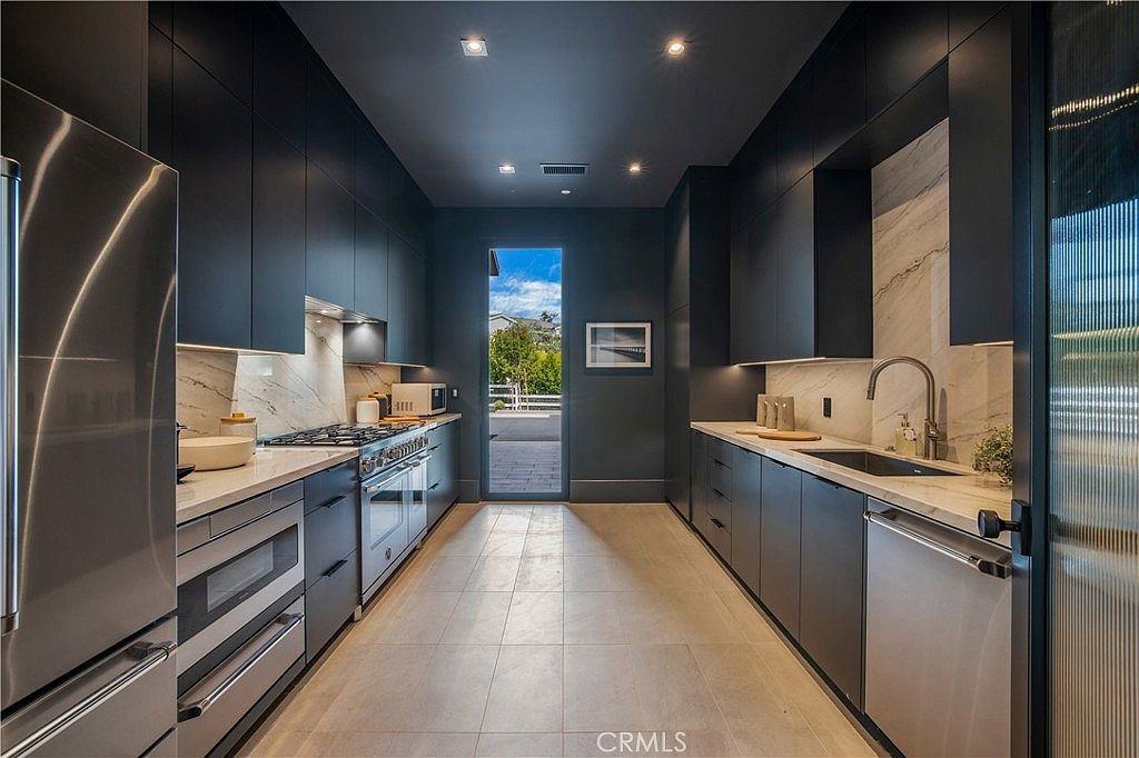This sleek, modern galley-style kitchen features dark, matte cabinetry contrasted by light marble countertops and backsplashes. The space is equipped with high-end stainless steel appliances, including a gas range and a built-in dishwasher, and is illuminated by recessed ceiling lights. A glass-paned door at the end of the corridor provides a bright, natural view of the outdoor area, creating a sophisticated and clean aesthetic.