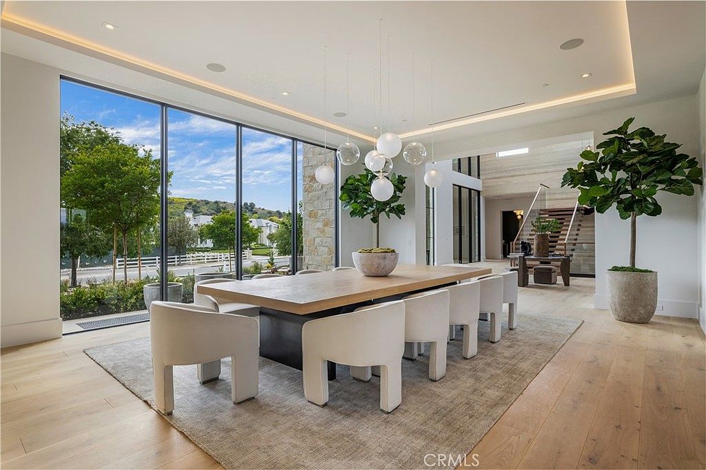 This elegant dining room features a large, light-wood table surrounded by modern, cream-colored upholstered chairs, set atop a textured area rug. A striking cluster of spherical pendant lights hangs above the table, while floor-to-ceiling windows provide an expansive view of the outdoors and fill the space with natural light. The room's minimalist design, complemented by a potted fiddle leaf fig and a view of a grand staircase in the background, creates a sophisticated and airy atmosphere.