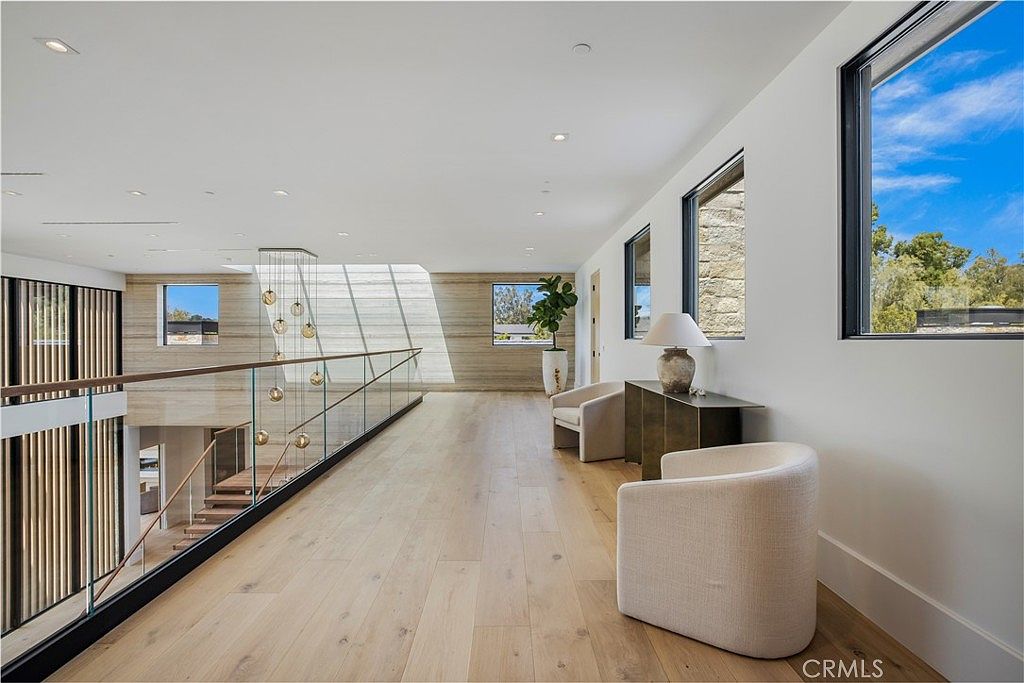This elegant, sun-drenched hallway features light-toned hardwood flooring and a sleek glass railing overlooking the staircase below. The space is accented by a modern chandelier, minimalist armchairs, and large windows that provide abundant natural light and views of the surrounding landscape. The overall aesthetic is sophisticated, airy, and contemporary, emphasizing clean lines and high-end finishes.