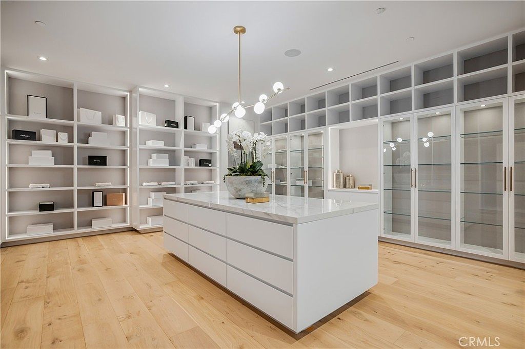 This luxurious, expansive walk-in closet features custom white cabinetry with a mix of open shelving and glass-fronted doors, providing ample storage and display space. A large, marble-topped center island serves as a focal point, complemented by a modern, multi-bulb chandelier and light-toned hardwood flooring. The room is bright and meticulously organized, offering a sophisticated and high-end aesthetic suitable for a primary suite.