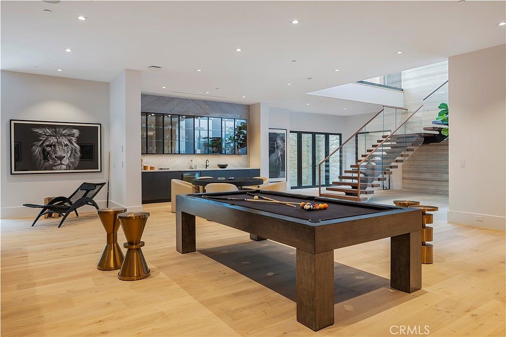 This expansive, modern basement recreation area features a prominent pool table as the central focal point, set against a backdrop of light hardwood flooring and neutral walls. The space includes a sophisticated wet bar with glass-front cabinetry and a striking floating staircase with glass railings that leads to the upper level. The room is decorated with contemporary art, including a large lion portrait, creating a luxurious and inviting atmosphere for entertainment.
