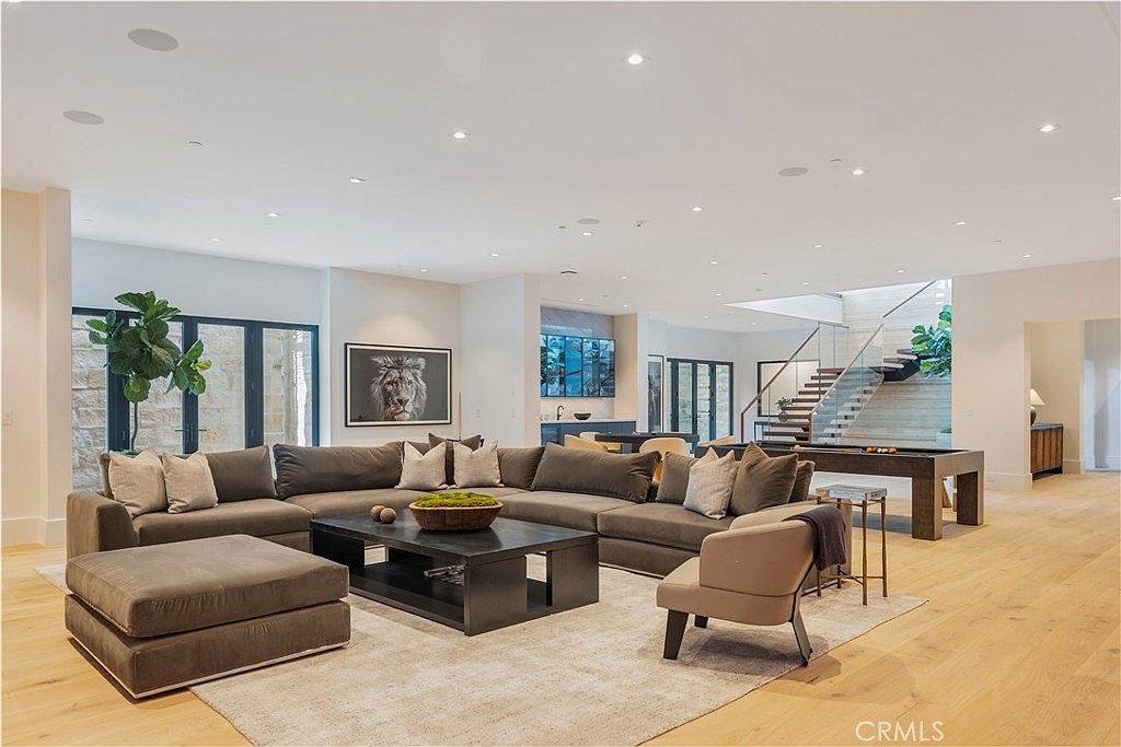This expansive, open-concept living room features a large, plush sectional sofa arranged around a dark wood coffee table, creating a sophisticated and inviting atmosphere. The space is highlighted by light hardwood flooring, recessed lighting, and a striking staircase in the background, all contributing to a modern and airy aesthetic. A large framed lion portrait serves as a bold focal point, while the seamless transition to the dining and stair areas emphasizes the home's spacious, high-end design.