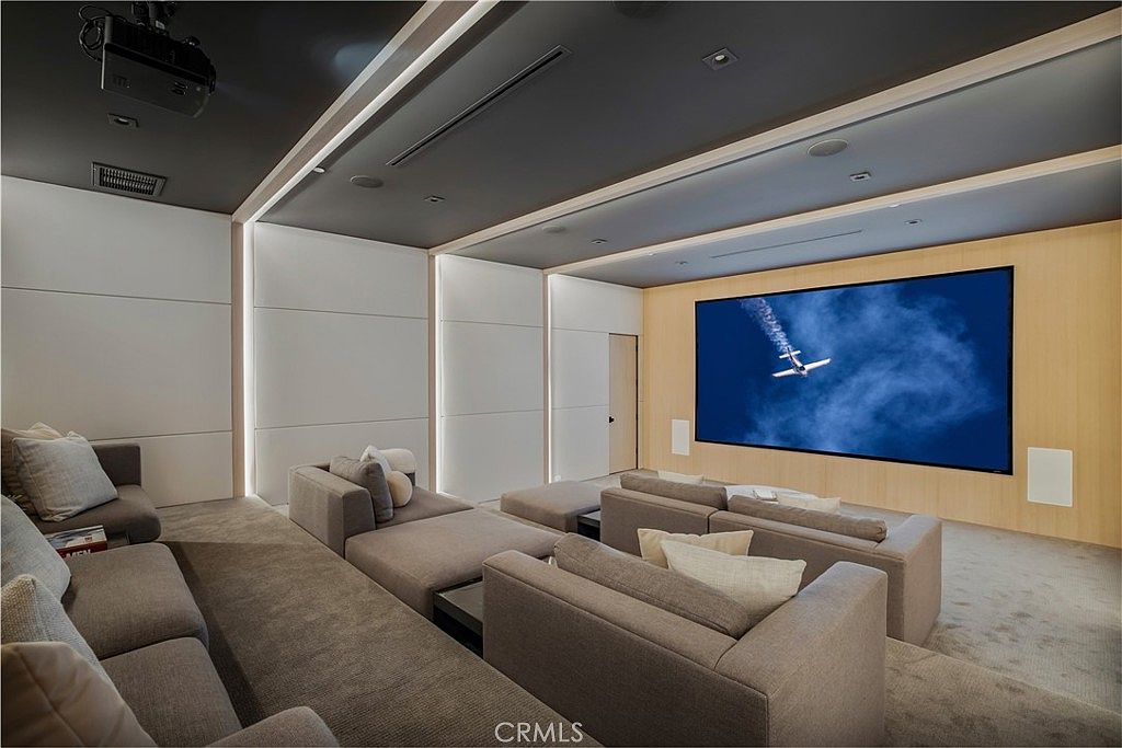 This image showcases a luxurious, modern home theater room designed for an immersive cinematic experience. The space features tiered plush gray seating, a large wall-mounted projection screen displaying an airplane, and sophisticated recessed lighting integrated into the ceiling and wall panels. The minimalist aesthetic, combined with warm wood accents and neutral tones, creates a high-end, comfortable atmosphere perfect for entertainment.