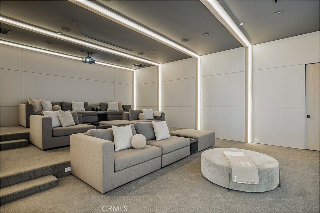 This image showcases a luxurious, tiered home theater room designed for ultimate comfort and cinematic viewing. The space features plush, light gray sectional seating arranged on two levels, complemented by a large, circular ottoman and minimalist, modern wall paneling with integrated LED accent lighting. The room's neutral color palette and clean lines create a sophisticated, high-end atmosphere perfect for private entertainment.