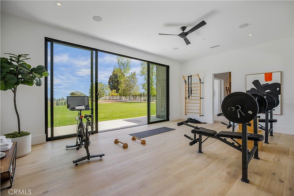 This bright and airy home gym features light-toned hardwood flooring and expansive floor-to-ceiling sliding glass doors that offer a seamless transition to the lush green backyard. The space is well-equipped with a stationary exercise bike, a weight bench with a barbell, and a wall-mounted wooden stall bar, creating a professional yet inviting workout environment. A large abstract painting adds a touch of modern art to the minimalist white walls, while a potted plant brings a refreshing natural element to the room.