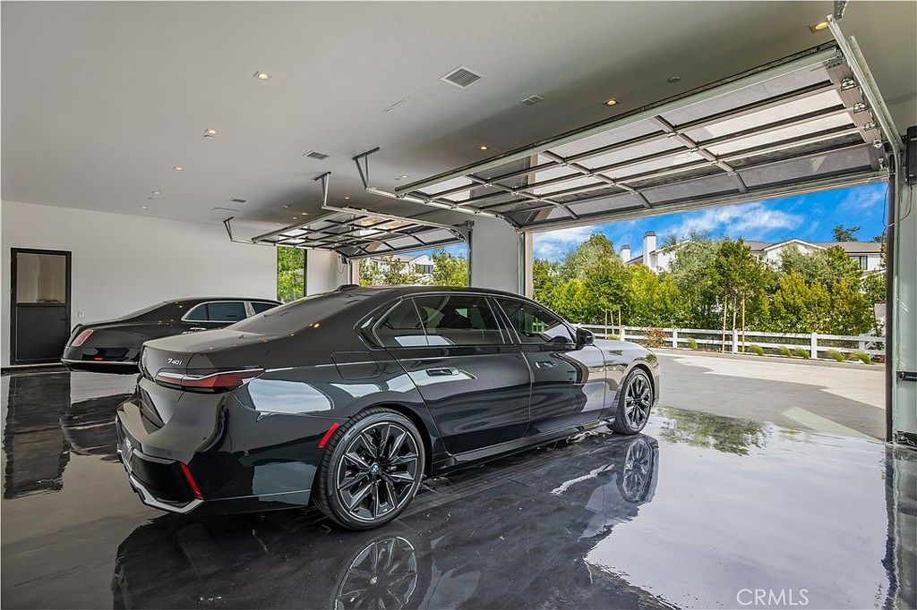 This image captures a sleek, modern garage interior featuring a high-gloss, reflective floor that showcases two luxury black sedans. The space is illuminated by recessed lighting and features a large, open glass-paneled garage door that provides a seamless transition to the outdoor landscape. The overall aesthetic is sophisticated and minimalist, emphasizing a high-end lifestyle.