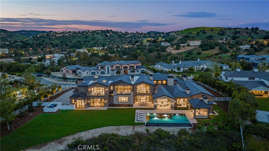 This stunning aerial view captures a sprawling, modern luxury estate at dusk, showcasing its expansive architectural design and seamless indoor-outdoor flow. The property features a large, illuminated swimming pool, a manicured lawn, and a covered patio area, all set against a backdrop of rolling hills and a twilight sky. The home's contemporary aesthetic is highlighted by its multi-level structure, dark rooflines, and large glass windows that glow warmly from within.