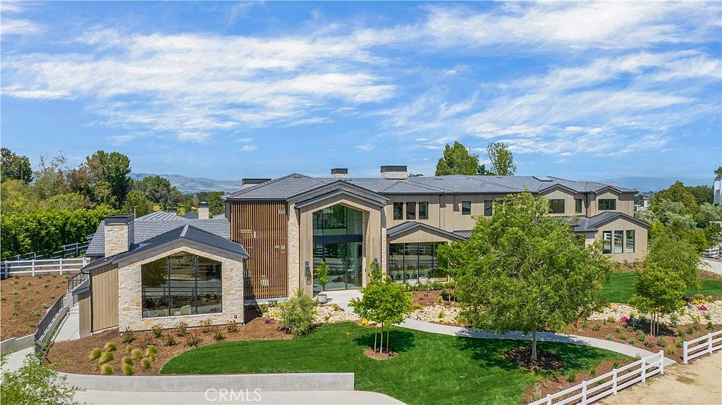 This stunning modern estate features a sophisticated blend of stone and wood siding, characterized by its grand, multi-gabled roofline and expansive glass windows. The property is set against a backdrop of lush green trees and a clear blue sky, with a meticulously landscaped lawn and a white rail fence framing the foreground. The architectural design exudes luxury and contemporary elegance, making it a standout residential property.