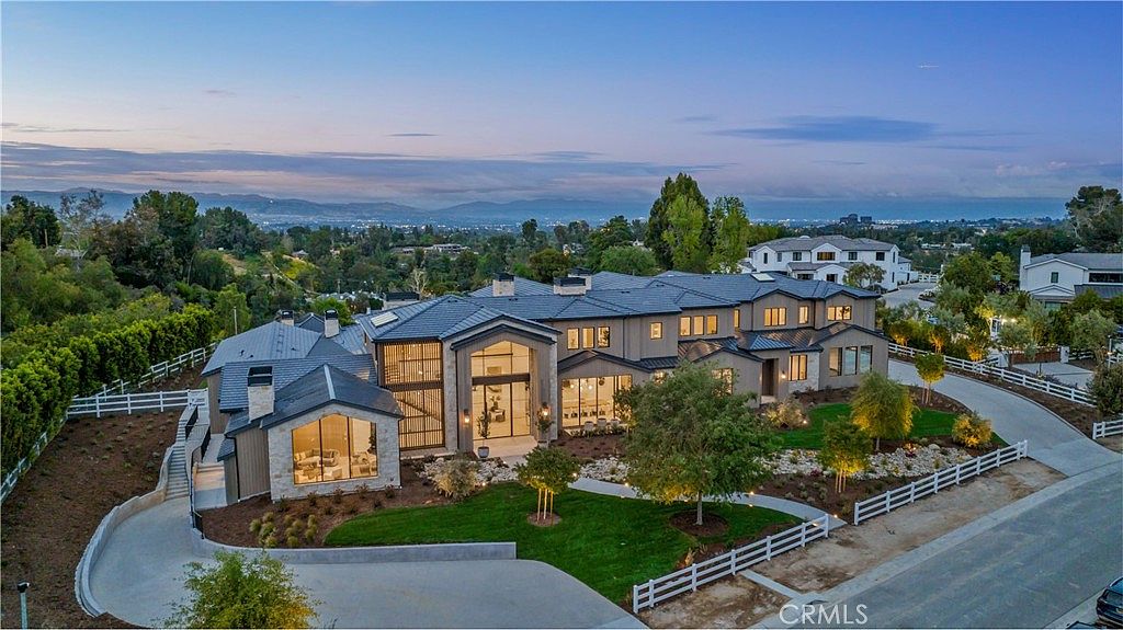 This stunning aerial view captures a sprawling, modern luxury estate set against a picturesque mountainous backdrop during the golden hour. The home features a sophisticated dark-tiled roof, stone and stucco exterior, and expansive glass windows that create a seamless blend of indoor and outdoor living. A manicured lawn, white split-rail fencing, and a winding driveway complete the elegant, high-end curb appeal of this property.