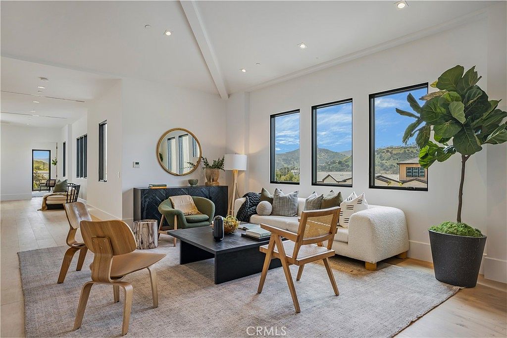 This bright and airy living room features a contemporary design with high vaulted ceilings and large windows that frame scenic mountain views. The space is furnished with a plush white sofa, a dark wood coffee table, and stylish wooden accent chairs, all anchored by a large neutral area rug. A sleek black console table with a circular mirror adds a sophisticated touch, creating an inviting and elegant atmosphere.