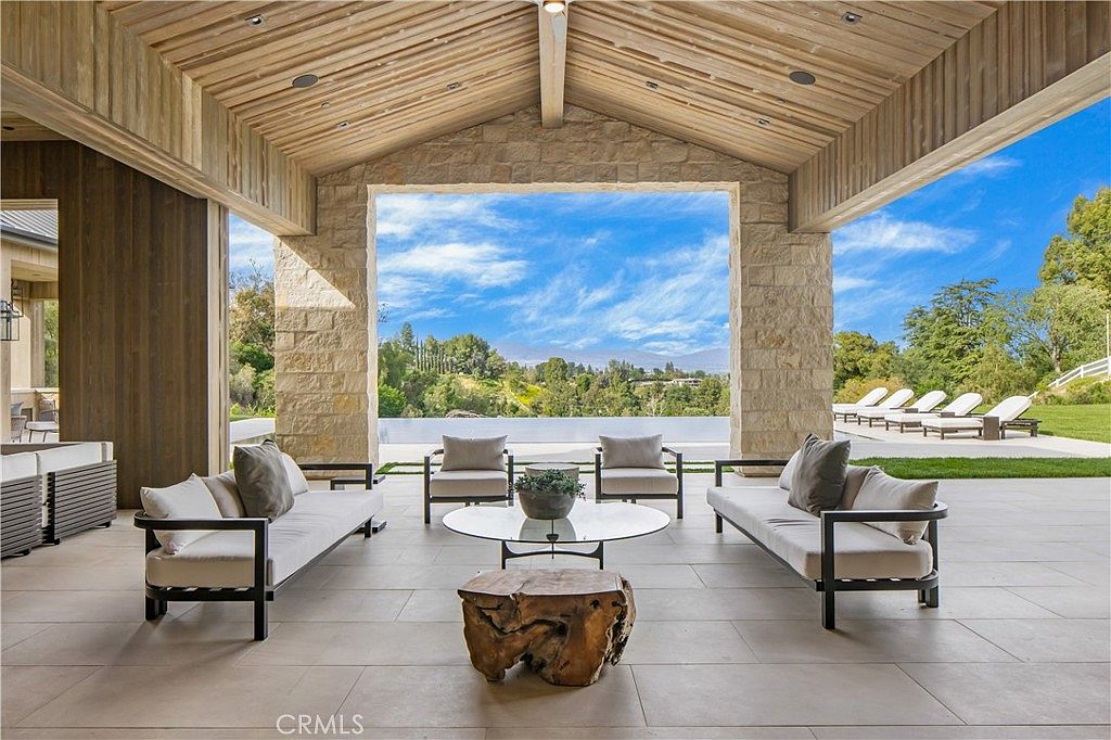 This luxurious covered patio features a high, wood-paneled vaulted ceiling and stone pillars that frame a stunning panoramic view of the surrounding hills and a sleek infinity pool. The space is furnished with two modern sofas, two armchairs, a glass-topped coffee table, and a rustic wooden stump accent piece, creating an elegant outdoor living area. The open-air design seamlessly blends indoor comfort with the natural beauty of the landscape, offering a serene and sophisticated atmosphere.