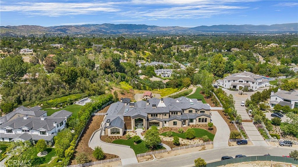 This high-angle aerial perspective showcases a sprawling, modern luxury estate nestled within a lush, hilly landscape. The property features a large, multi-gabled residence with a grey roof, extensive paved driveways, and manicured green lawns, all set against a backdrop of rolling mountains and suburban greenery. The composition emphasizes the home's impressive scale and its integration into a serene, upscale residential community.