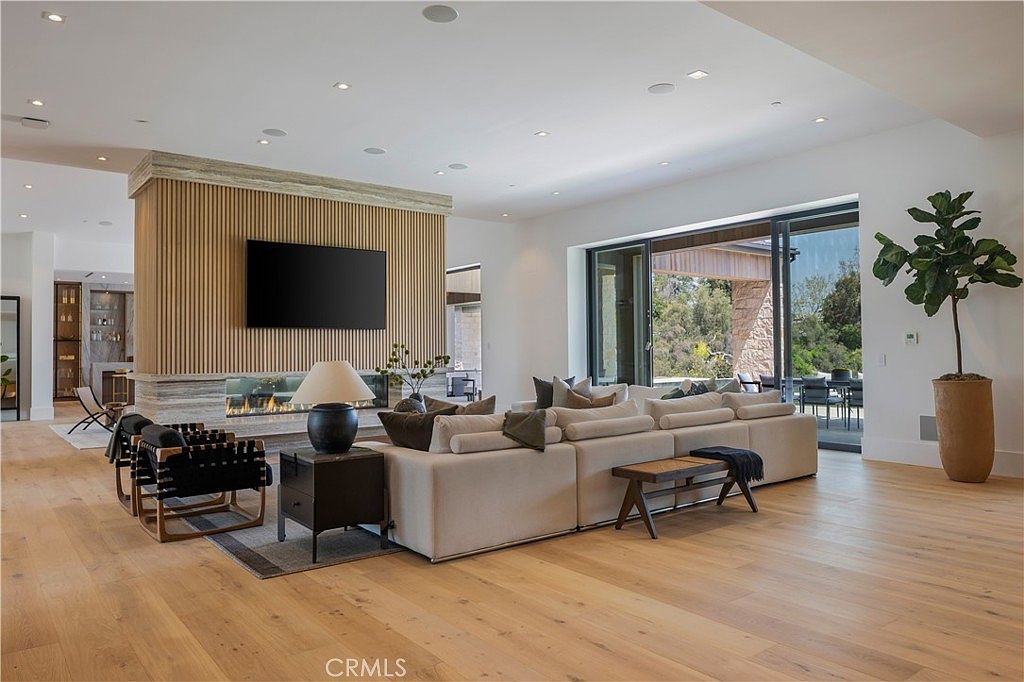 This spacious, open-concept living room features a striking floor-to-ceiling wood-slat accent wall with a built-in fireplace and television. A large, plush sectional sofa anchors the room, which is bathed in natural light from expansive sliding glass doors that open to an outdoor patio. The minimalist design, light hardwood flooring, and neutral color palette create an airy, sophisticated, and inviting atmosphere.