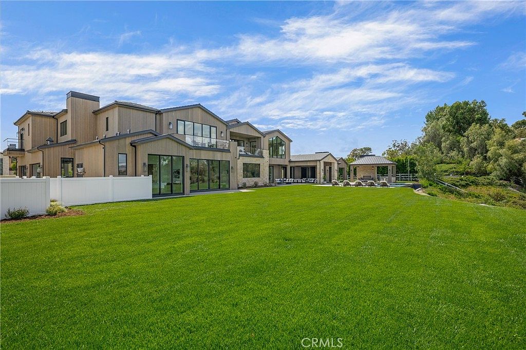This expansive rear view of a luxury estate showcases a modern architectural design with light-toned wood siding and large glass windows. The property features a vast, meticulously manicured green lawn that leads to a covered outdoor patio area and a swimming pool in the distance. The bright blue sky with wispy clouds provides a serene backdrop, emphasizing the home's grand scale and resort-like atmosphere.