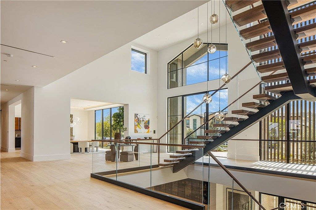 This striking interior view showcases a grand, modern staircase with floating wooden treads and a sleek glass railing, serving as the architectural centerpiece of the home. The open-concept design features soaring ceilings, expansive windows that flood the space with natural light, and a seamless transition to an adjacent dining area. The minimalist aesthetic, characterized by clean white walls and light hardwood flooring, creates an airy, sophisticated, and luxurious atmosphere.