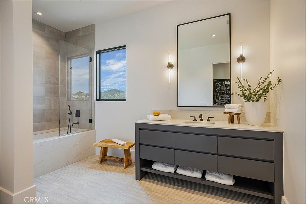 This modern primary bathroom features a sleek, dark gray vanity with a white countertop and a large, rectangular mirror flanked by contemporary wall sconces. To the left, a glass-enclosed shower and bathtub combination is finished with light-toned, large-format tiles, while a rustic wooden stool adds a touch of organic warmth to the space. The room is bright and airy, with a window providing natural light and a view of the surrounding landscape.