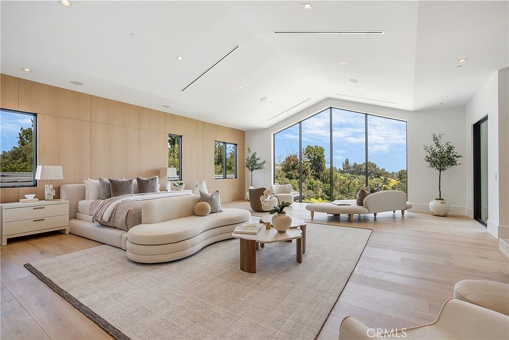 This expansive primary bedroom features a sophisticated, neutral color palette with light wood paneling and high, vaulted ceilings. The room is anchored by a large, curved sofa at the foot of the bed and floor-to-ceiling windows that provide a panoramic view of the surrounding landscape. The minimalist design, combined with high-end furnishings and a spacious layout, creates an atmosphere of serene luxury.