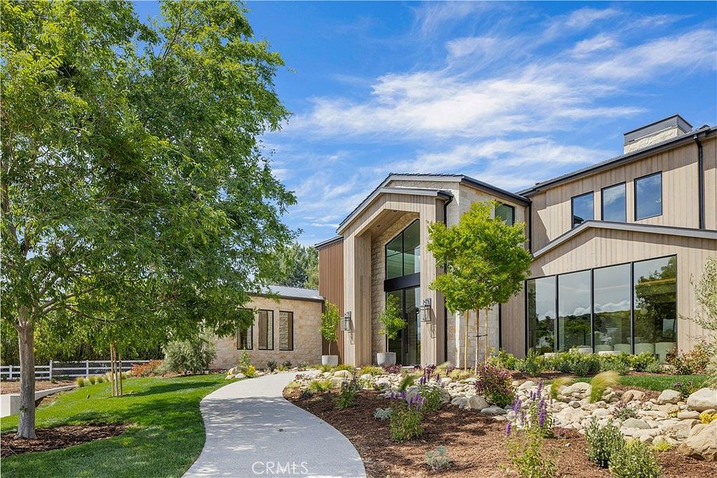 This stunning modern estate features a striking architectural design with a blend of natural stone, warm wood siding, and expansive floor-to-ceiling glass windows. A curved concrete pathway leads visitors through a meticulously landscaped front yard, complete with decorative rock beds and lush greenery. The bright, sunny atmosphere and clean lines create an inviting and sophisticated first impression of this luxury residence.