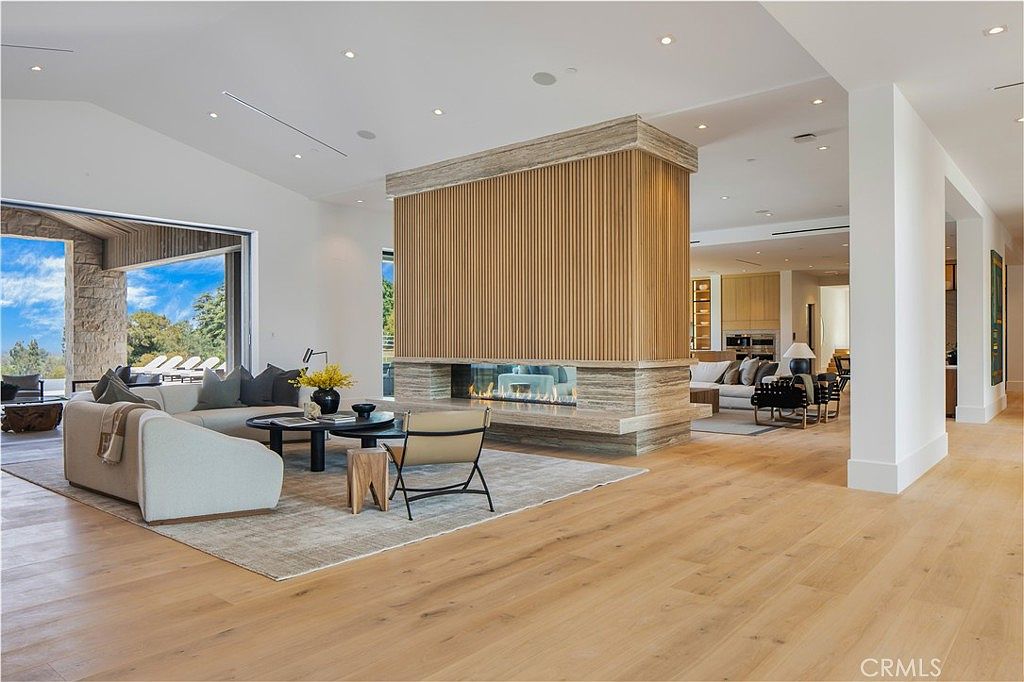 This expansive, open-concept living area features a striking central fireplace with vertical wood slat detailing and a stone hearth, serving as a sophisticated focal point. The room is bathed in natural light from large sliding glass doors that open to an outdoor patio, highlighting the light-toned hardwood flooring and minimalist, contemporary furnishings. The space flows seamlessly into an adjacent seating area, creating an airy and luxurious atmosphere perfect for modern entertaining.