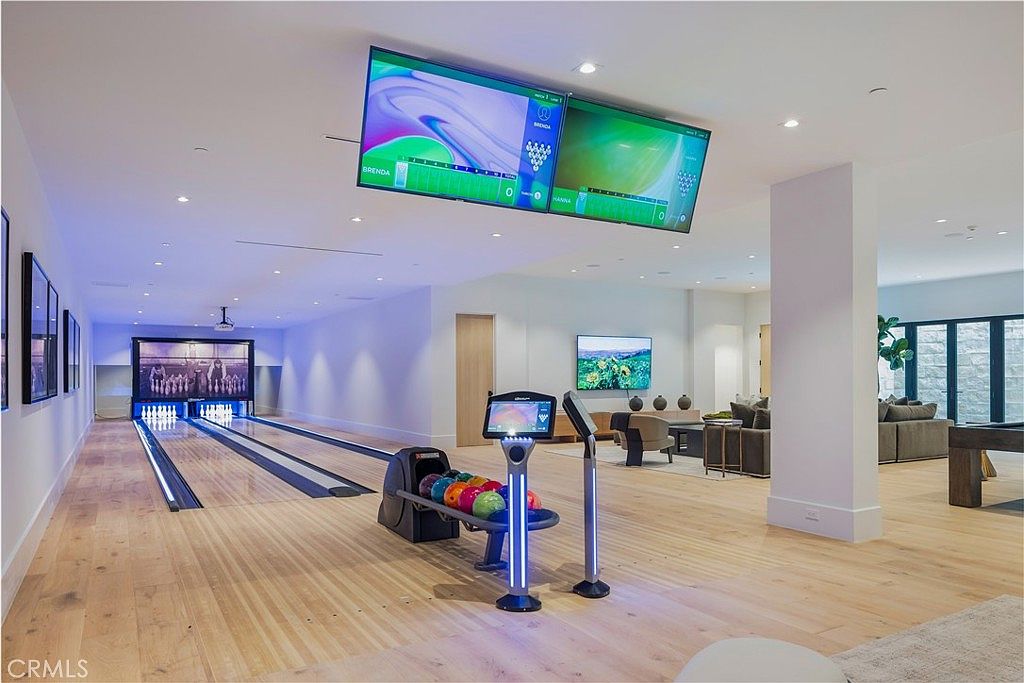This image showcases a luxurious private bowling alley integrated into a spacious, modern basement entertainment area. The room features light hardwood flooring, a professional-grade bowling lane with a digital scoring display, and a comfortable lounge area with plush seating in the background. The aesthetic is sleek and contemporary, designed for high-end recreation and social gatherings.