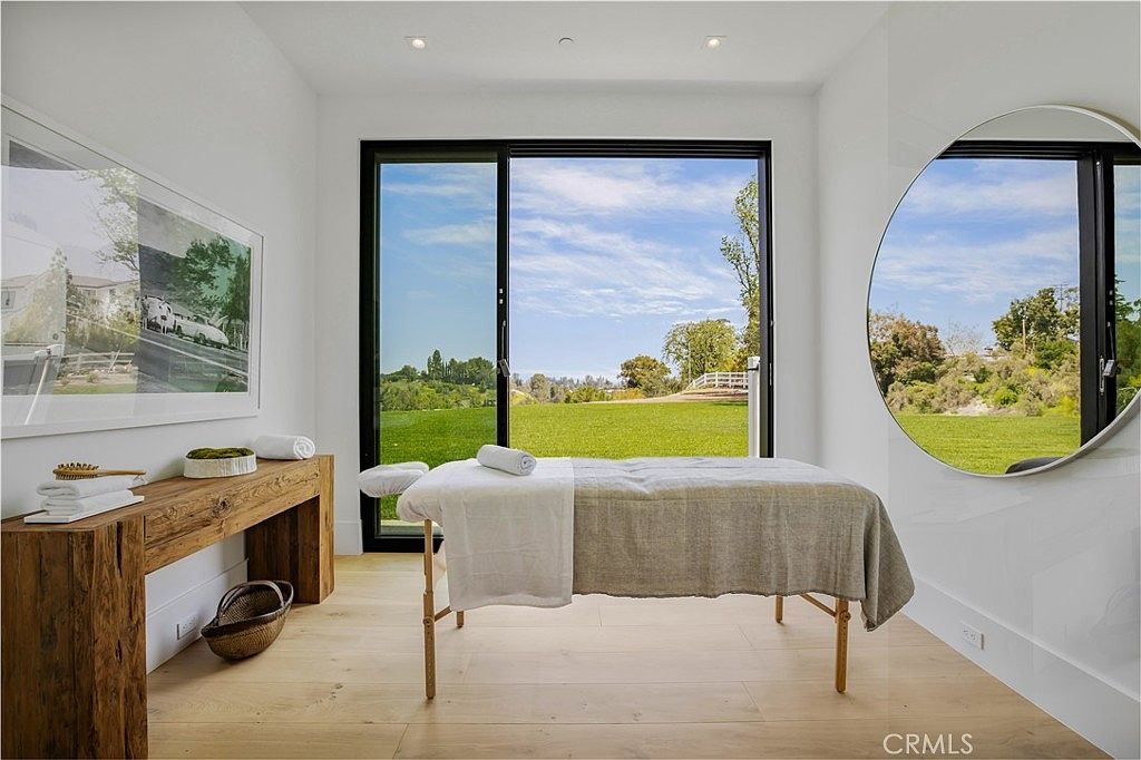 This serene spa or massage room features a minimalist, modern aesthetic with light wood flooring and crisp white walls. A massage table is centered in the room, positioned in front of a large floor-to-ceiling sliding glass door that offers a tranquil, expansive view of a lush green lawn and trees. The space is accented by a large circular mirror, a rustic wooden console table, and soft recessed lighting, creating a peaceful and luxurious atmosphere.