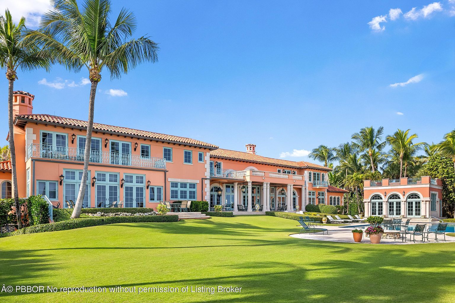 This image showcases the rear exterior of a large, luxurious estate with a sprawling green lawn. The house features a Mediterranean-inspired design with a terracotta roof, peach-colored walls, and blue-trimmed windows and balconies. A pool and pool house are visible in the background, surrounded by lush palm trees, creating a resort-like atmosphere.