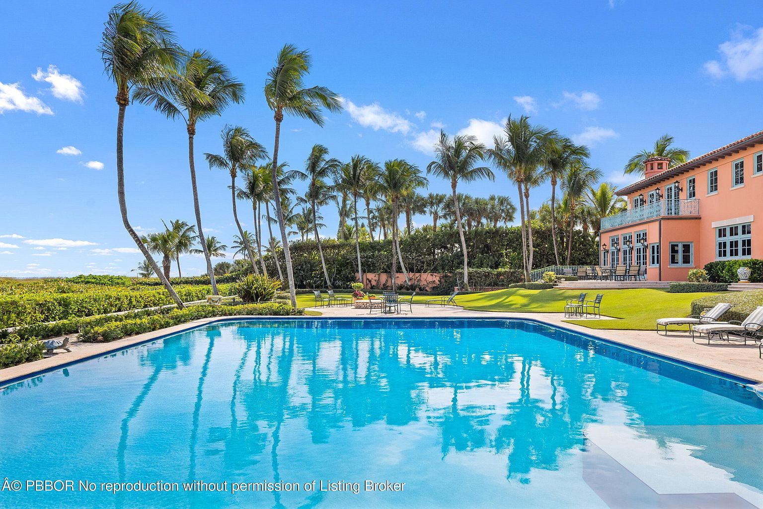 This image showcases a luxurious pool area with a large, inviting pool reflecting the surrounding palm trees and blue sky. The pool is bordered by a well-manicured lawn, leading to a grand, peach-colored house with a balcony. The scene exudes a sense of tropical elegance and relaxation, perfect for high-end real estate appeal.