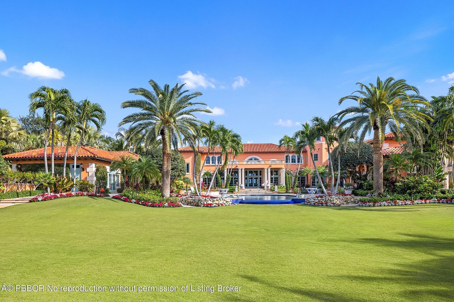 This is a stunning front view of a luxurious estate, showcasing a grand residence with a symmetrical facade, terracotta roof, and elegant columns. The property features meticulously manicured lawns, vibrant flower beds, and mature palm trees, creating a tropical paradise. A circular pool adds a touch of opulence, enhancing the overall curb appeal and highlighting the property's high-end features.