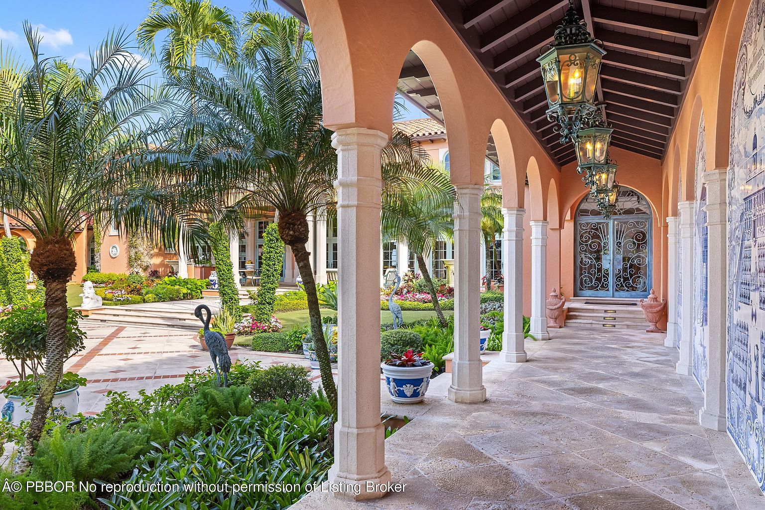 This image showcases a beautifully landscaped yard and garden, viewed from a covered walkway with arched columns. The garden features lush greenery, palm trees, statues, and meticulously arranged flower beds. The walkway is adorned with hanging lanterns and decorative tile work, creating an elegant and inviting outdoor space.