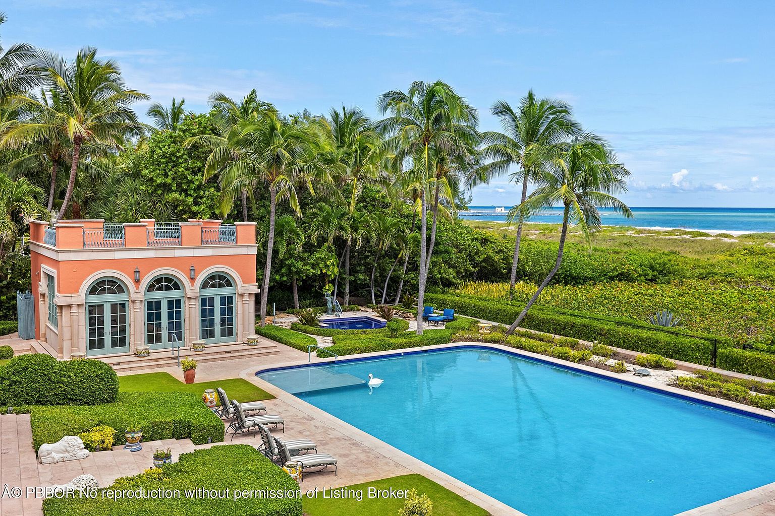 This image showcases a luxurious outdoor pool area with a large, inviting pool featuring a swan float. The pool is surrounded by meticulously manicured lawns, hedges, and palm trees, creating a tropical oasis. A charming pool house with arched windows and a balcony adds architectural interest, while the ocean and beach are visible in the background, emphasizing the property's prime location.