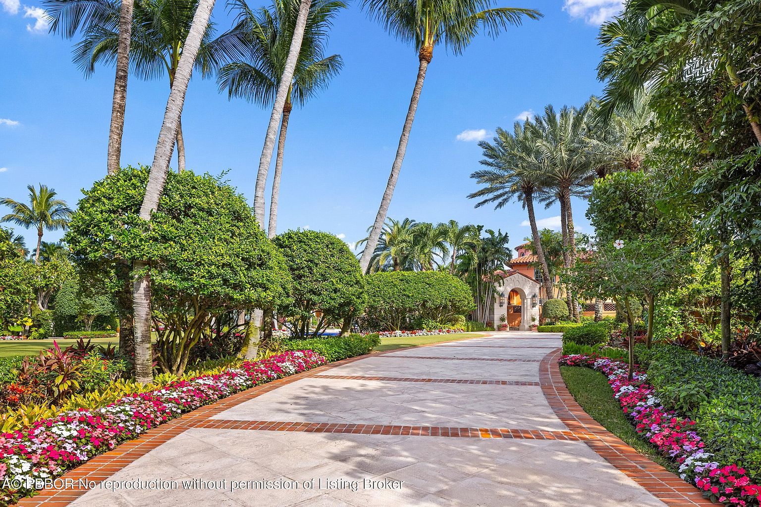 This image showcases a grand entryway leading to a luxurious estate. A meticulously designed driveway, bordered by vibrant flowerbeds and lush greenery, guides the eye towards an elegant arched entrance. Tall palm trees frame the scene, adding a touch of tropical sophistication and emphasizing the property's upscale appeal.