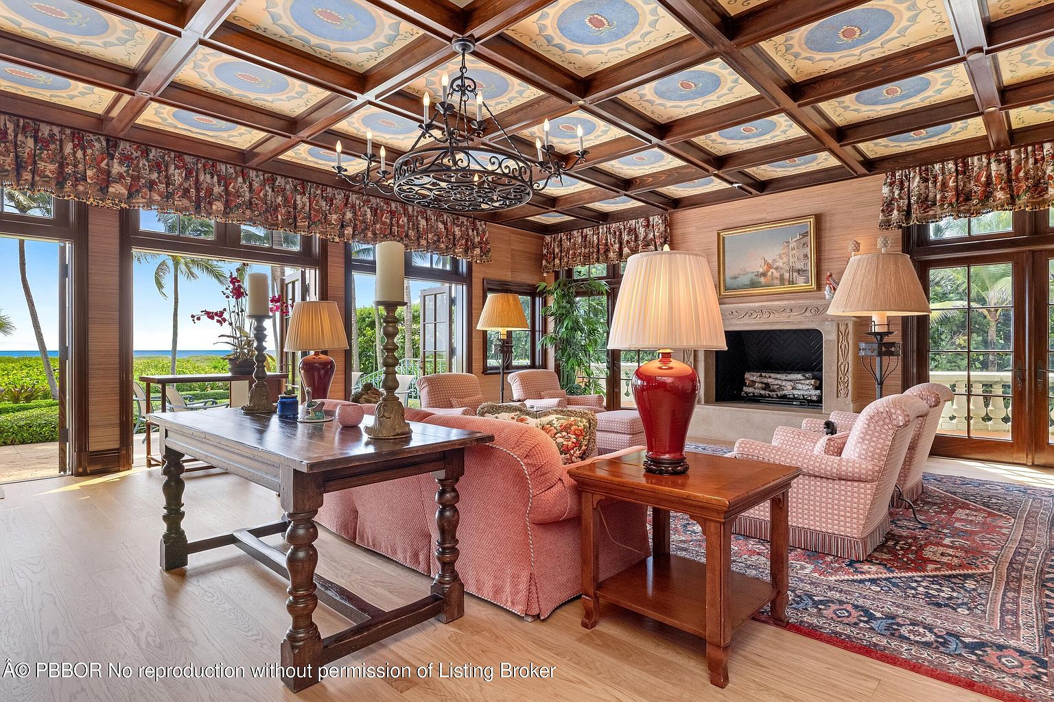 This is an interior shot of a luxurious living room featuring a unique coffered ceiling with painted details, a large chandelier, and multiple seating areas. The room is filled with natural light from the large windows offering ocean views, and a fireplace adds a cozy touch. The overall style is elegant and sophisticated, creating a warm and inviting atmosphere.
