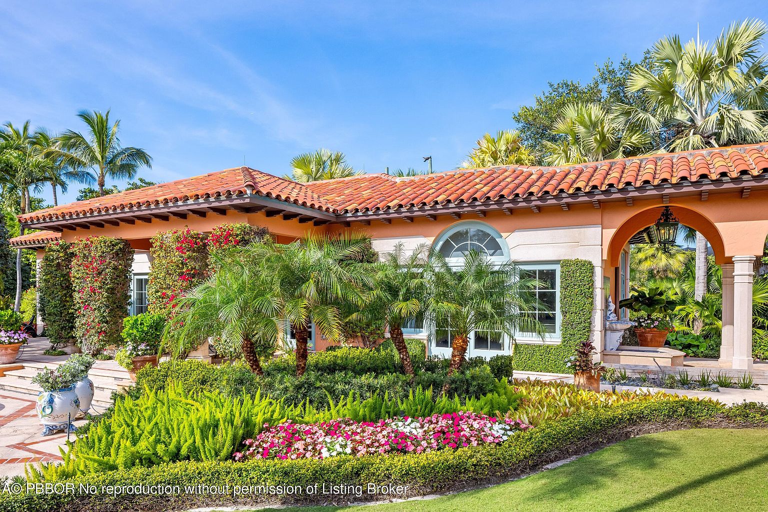 This is a front view of a luxurious Mediterranean-style home, showcasing a terracotta tile roof, stucco walls, and meticulously landscaped gardens. The property features an arched entryway, lush greenery, and vibrant flowerbeds, creating an inviting and elegant curb appeal. The clear blue sky enhances the overall impression of a well-maintained and desirable residence.