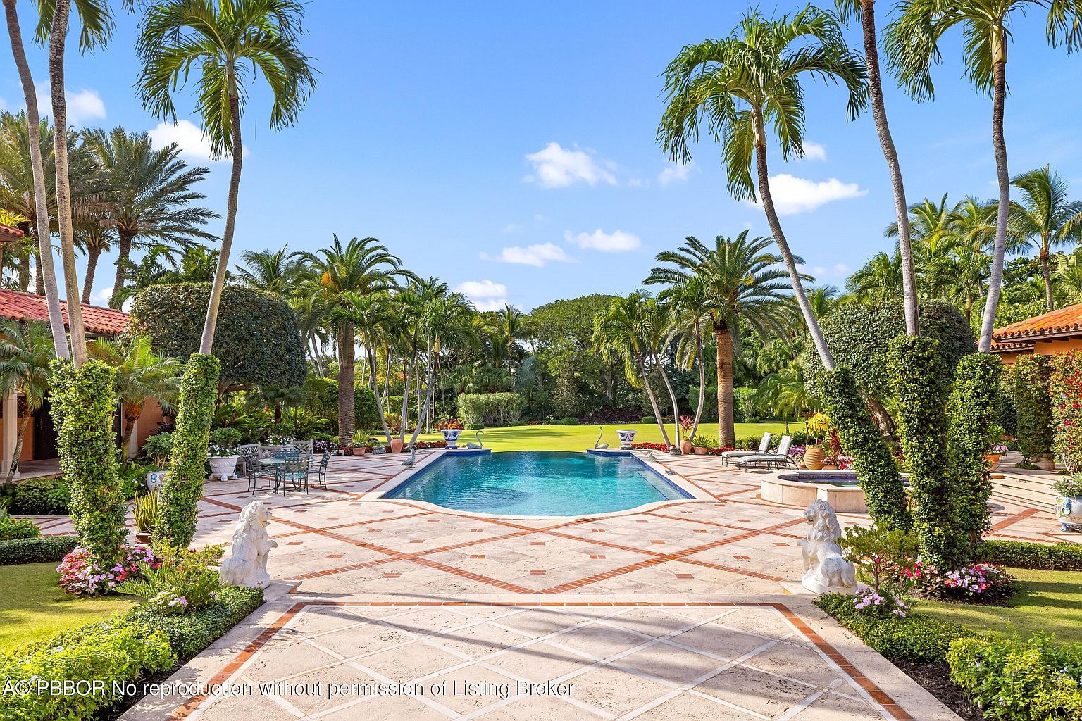 This image showcases a luxurious backyard featuring a beautifully designed pool surrounded by a patterned tile patio. Lush landscaping with palm trees and manicured gardens frame the pool area, creating a serene and private oasis. Statues flank the entrance to the pool area, adding a touch of elegance and sophistication to the outdoor space.