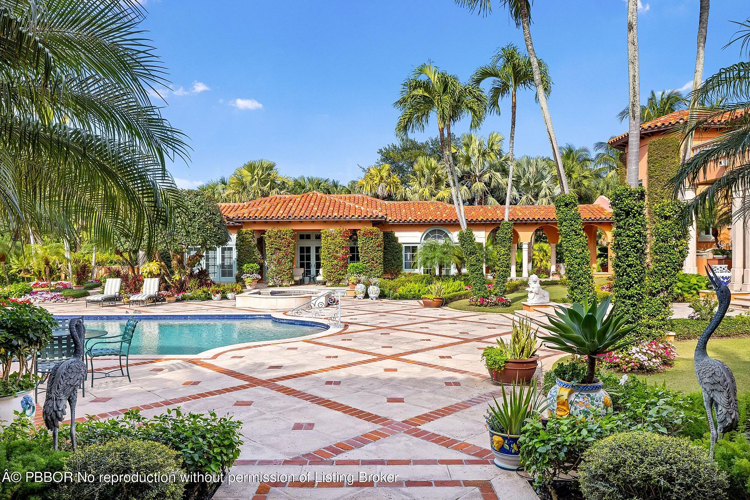 This image showcases a luxurious backyard featuring a swimming pool and spa area. The pool is surrounded by a patterned brick patio, lush landscaping, and mature palm trees. The architecture of the house is visible in the background, featuring a red tile roof and arched walkways, creating an elegant and inviting outdoor living space.