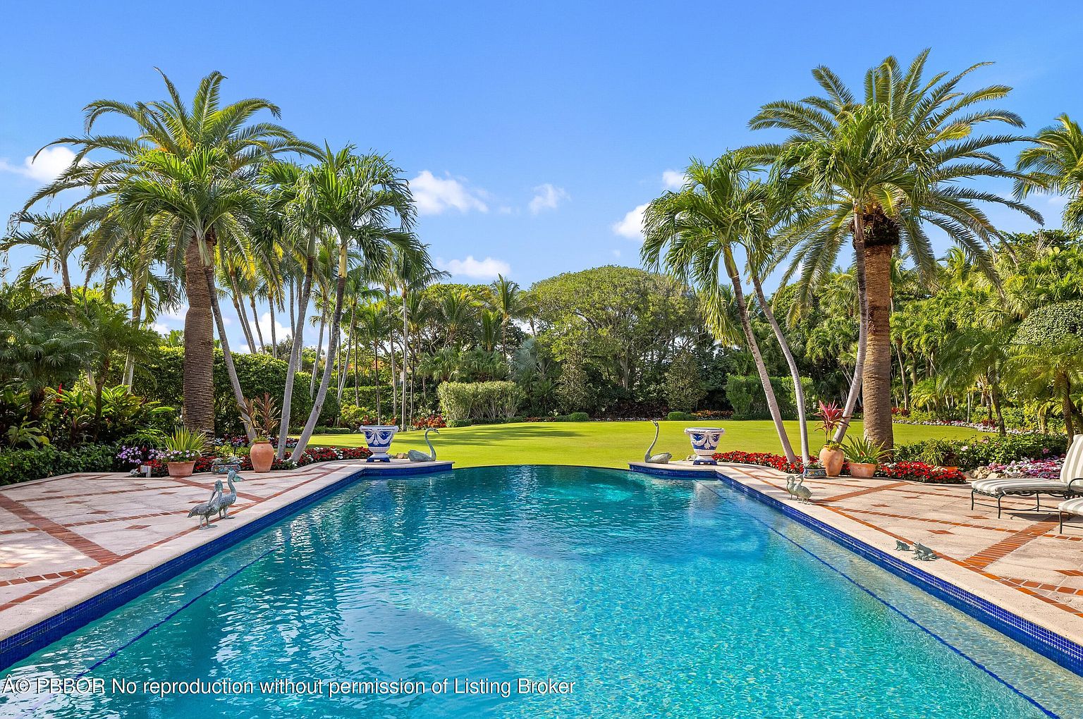 This image showcases a luxurious swimming pool surrounded by lush landscaping, including tall palm trees and manicured lawns. The pool's clear blue water invites relaxation, while decorative urns and swan statues add an elegant touch to the poolside area. The overall impression is one of a private, resort-like oasis.
