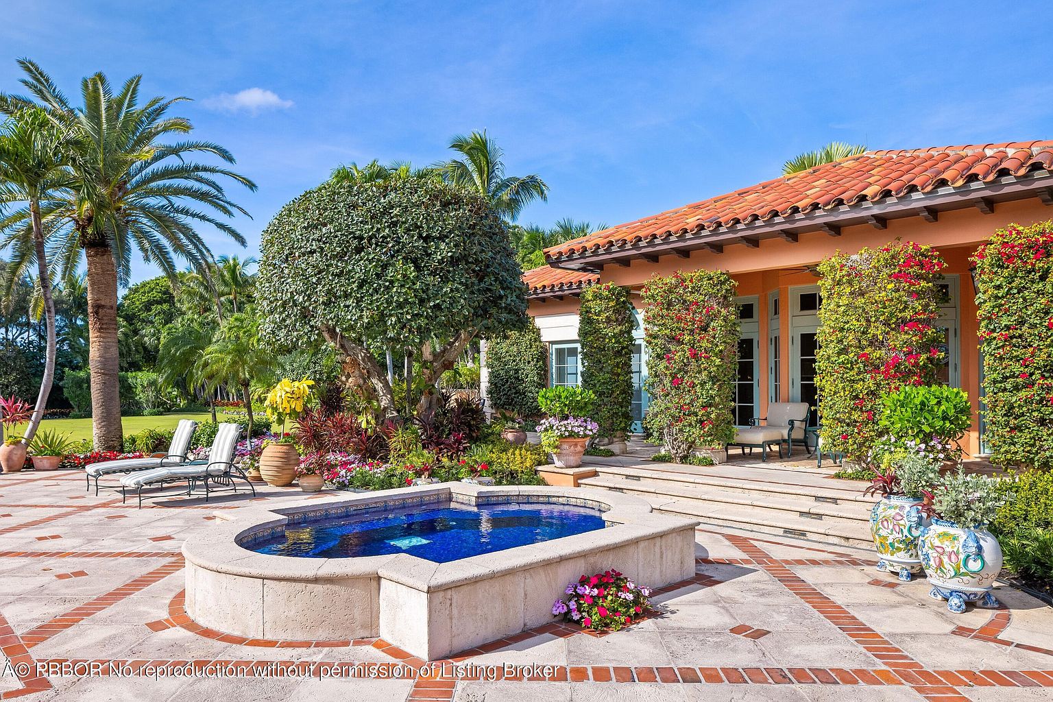 This image showcases a beautifully landscaped backyard featuring a tiled patio with a small, elegant water feature in the foreground. Lush greenery, including palm trees and manicured shrubs, surrounds the patio, leading to a Mediterranean-style house with a terracotta tile roof and vine-covered columns. The scene exudes a sense of luxury and tranquility, perfect for outdoor relaxation and entertaining.