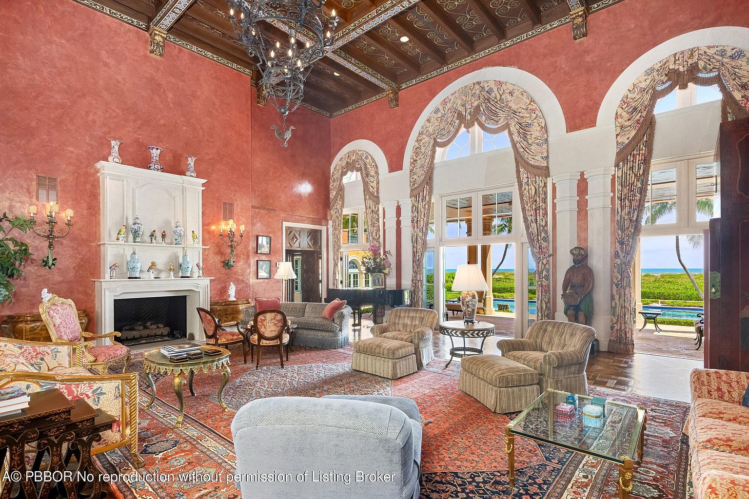 This is a grand living room featuring a high ceiling with intricate detailing, a large chandelier, and a fireplace with decorative elements. The room is furnished with ornate chairs, sofas, and tables, and a large rug covers the floor. Large arched windows offer a view of the exterior, creating a luxurious and inviting atmosphere.
