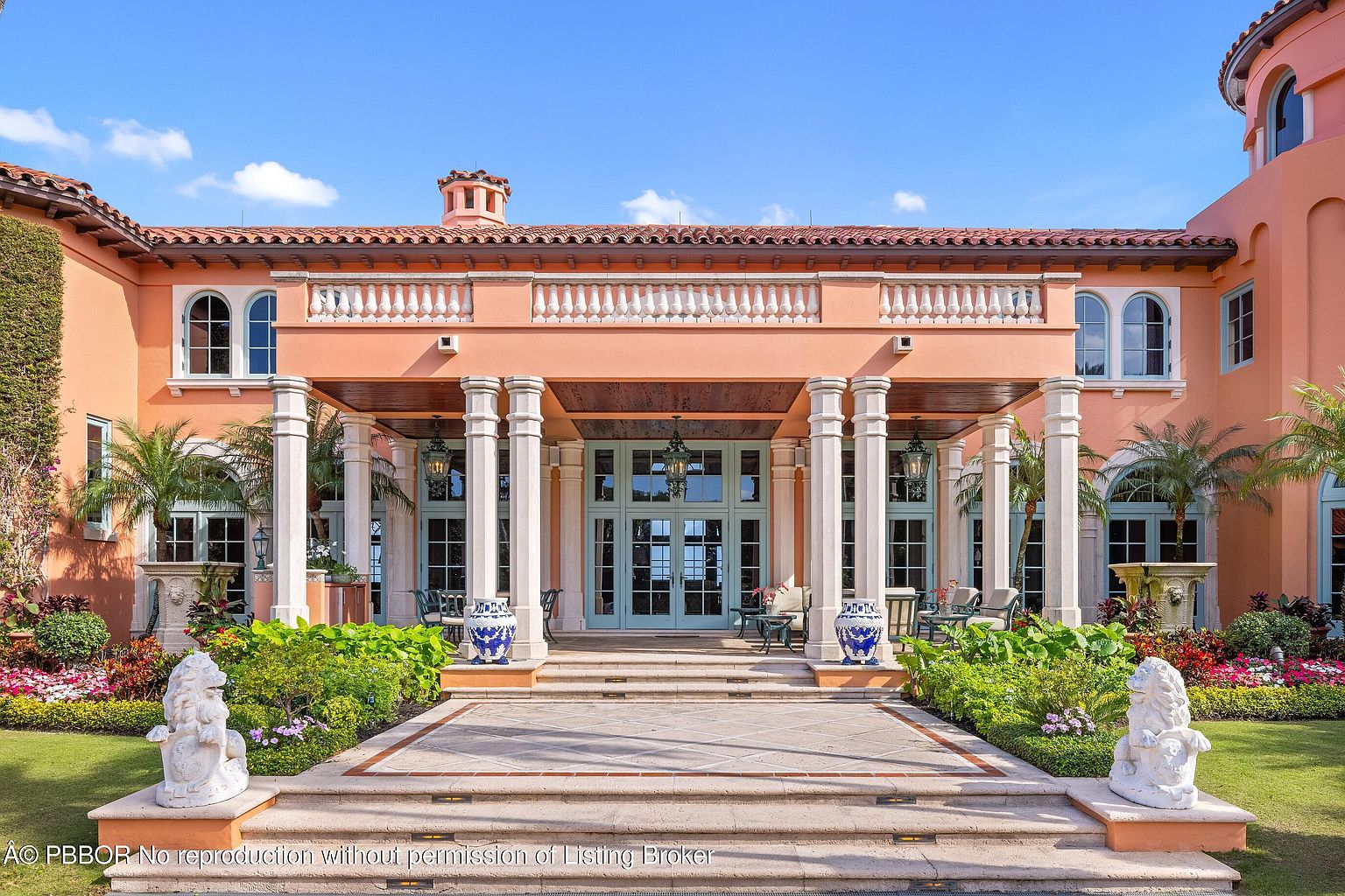 This is a grand entryway to a luxurious Mediterranean-style home. The entrance features a covered portico supported by large white columns, leading to double doors. The landscaping is meticulously maintained with manicured gardens, stone lion statues, and a tiled walkway, creating an inviting and impressive approach to the property.