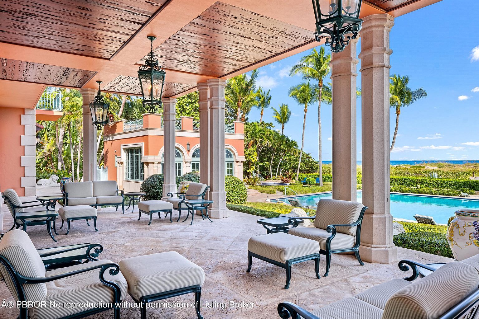 This image showcases a luxurious outdoor patio area with elegant seating arrangements under a covered structure supported by stately columns. The patio overlooks a beautifully landscaped yard with a swimming pool and glimpses of the ocean, creating a serene and upscale ambiance. The architectural details and high-end furnishings suggest a sophisticated and comfortable outdoor living space.