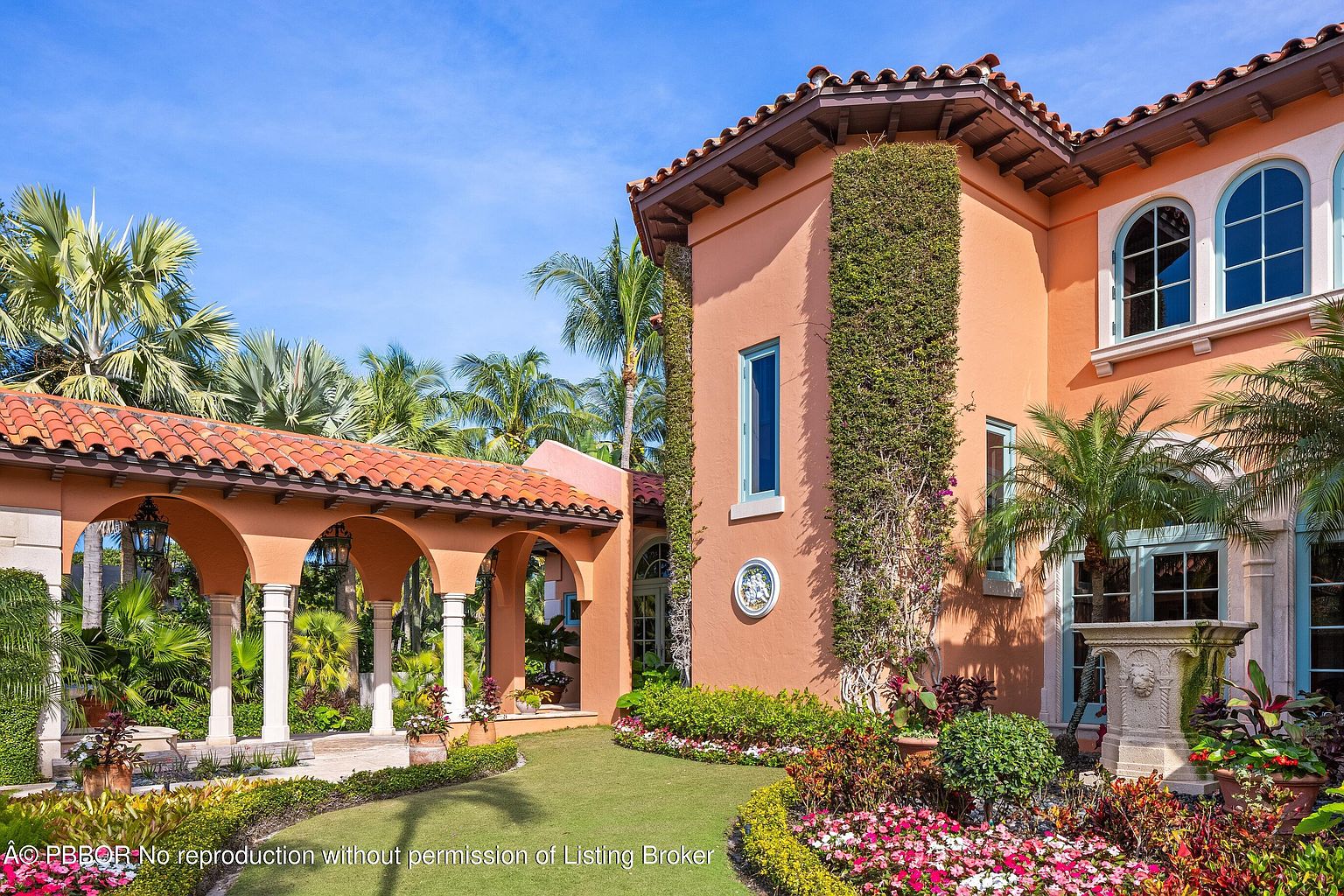 This image showcases the exterior of a luxurious Mediterranean-style home, emphasizing its meticulously landscaped yard and garden. The architecture features arched walkways with white columns, a terracotta tile roof, and walls adorned with climbing vines. The vibrant flowerbeds and lush greenery create an inviting and elegant atmosphere, highlighting the property's curb appeal and outdoor living spaces.