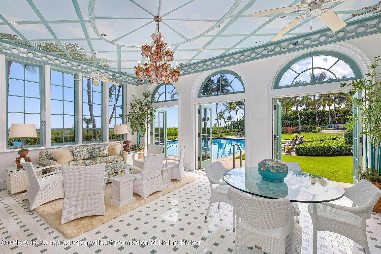 This sunroom features a bright and airy atmosphere with large windows offering views of the pool and garden. The room is furnished with white wicker furniture, a glass-top dining table, and a floral-patterned sofa, creating a relaxed and elegant space. The unique ceiling design and chandelier add a touch of sophistication.
