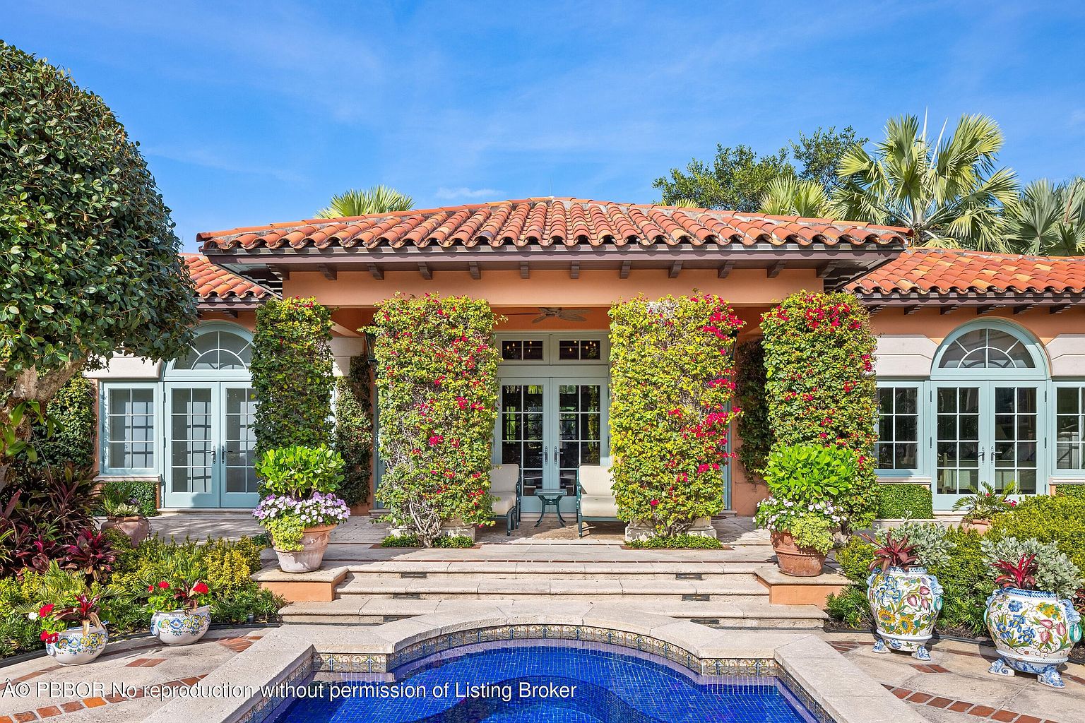 This is a front view of a luxurious Mediterranean-style home. The facade features a terracotta tile roof, light peach stucco walls, and arched windows with light blue frames. Lush greenery, including flowering vines on the pillars and potted plants, adds to the property's curb appeal, while a blue-tiled pool is visible in the foreground.